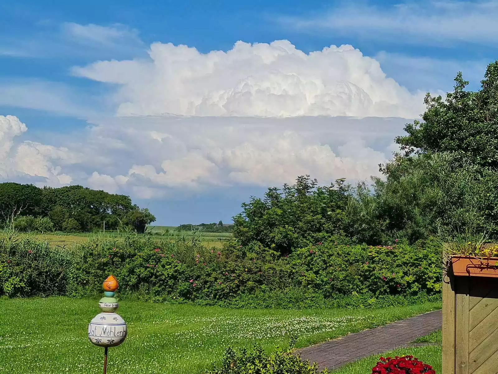 Garten des Dat Möhlenhus mit Blumen, Dekoration und weitem Blick über Felder und Wiesen unter nordischem Wolkenhimmel